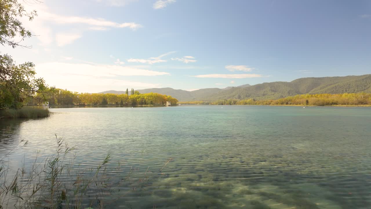 lake surrounded by lush greenery and autumn-colored trees In the distance, forested hills and mountains rise, creating a peaceful, picturesque scene that captures