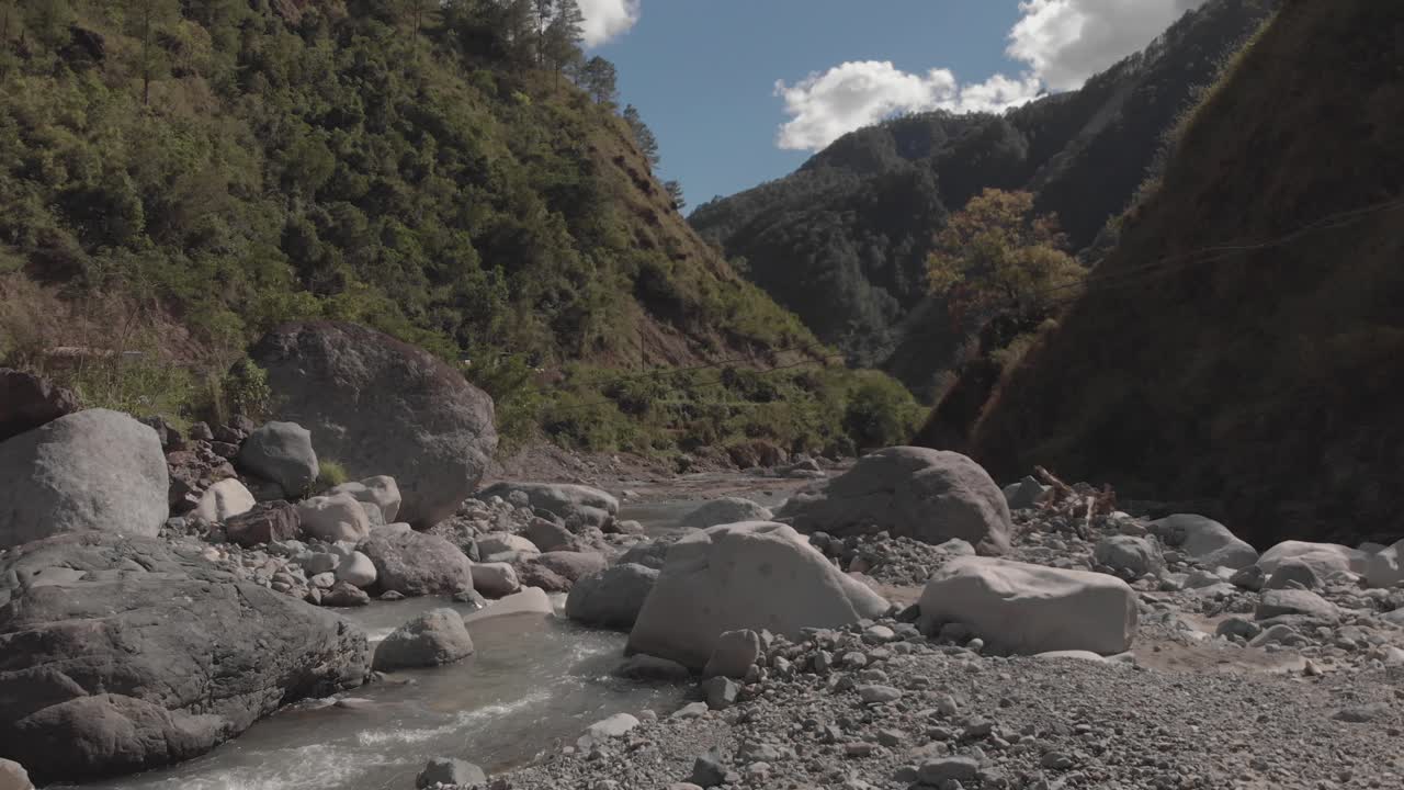 río rocoso que serpentea a través de montañas en el valle del cañón, agua que fluye acercándose a rocas grises, árboles verdes, cielo azul y nubes, antena lenta, avance suave, proximidad a cables eléctricos