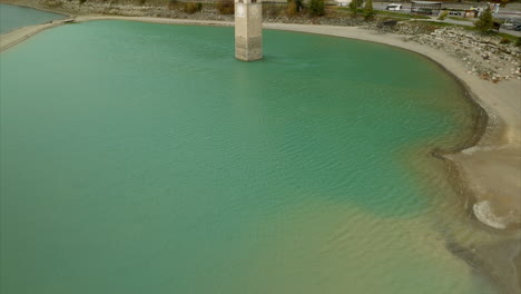 aerial tilt-up reveals sunken old bell tower in the middle of lake resia alongside a quaint village and coniferous forest during summer