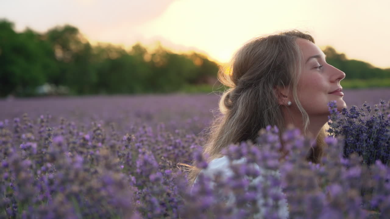 Woman in a white dress holding and smelling a bouquet of lavender in a field at sunset
