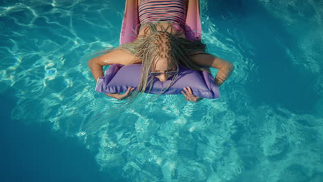 Top-view:-A-child-with-afro-pigtails-swims-on-an-inflatable-mattress-in-the-pool,-resting-and-enjoying-the-rest.