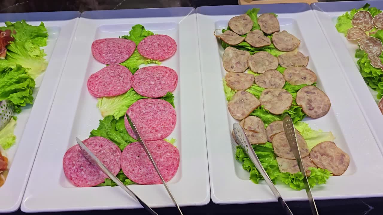 Right-to-left panning shot of assorted cold cuts on white trays with tongs