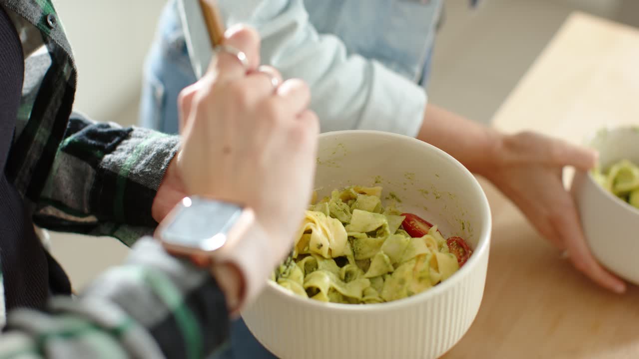 Two female friends stirring pasta in ceramic bowl on counter while friend bringing second bowl