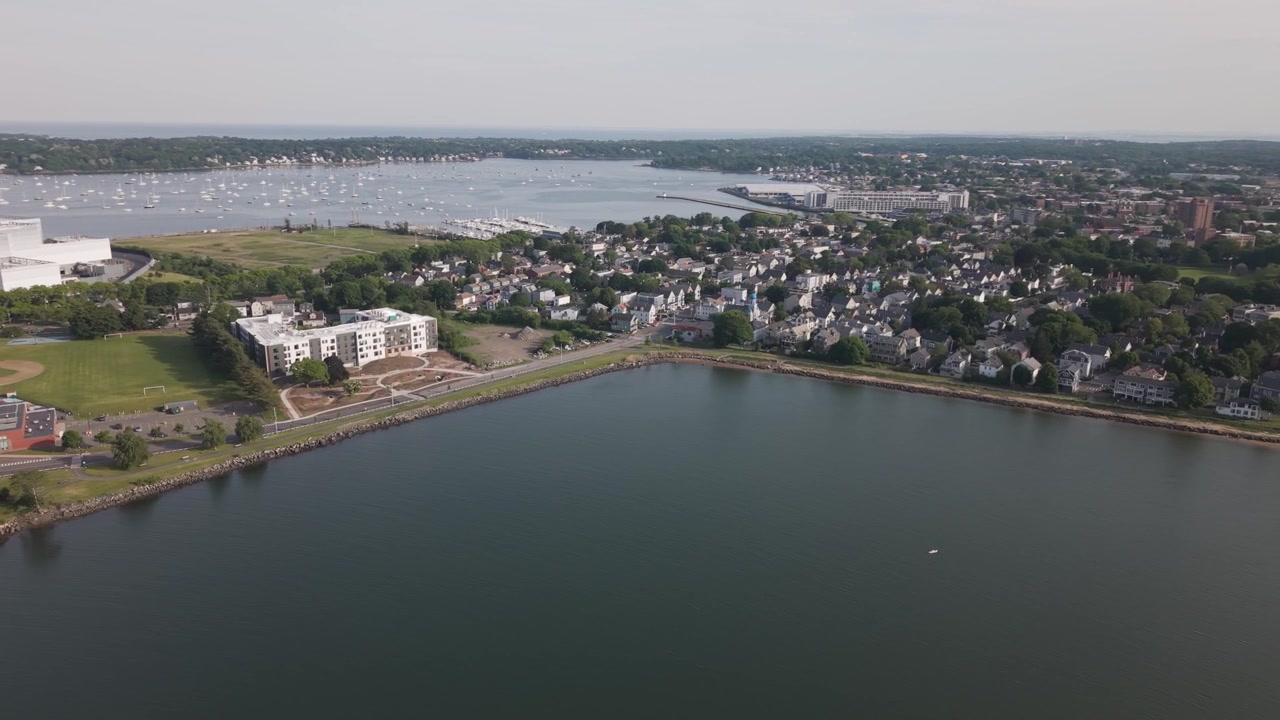 Complexo de apartamentos branco moderno ao lado de campo de futebol e estrada à beira-mar em Salem, Massachusetts, com telhados densos e marina distante se estendendo pela água calma do porto. Imagem aérea ampla de drone.