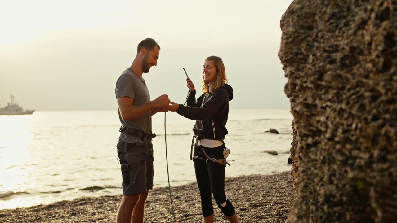 una chica rubia con el cabello rizado en ropa negra enseña a su novio moreno en una camiseta gris a atar nudos especiales para hacer belay mientras se dobla