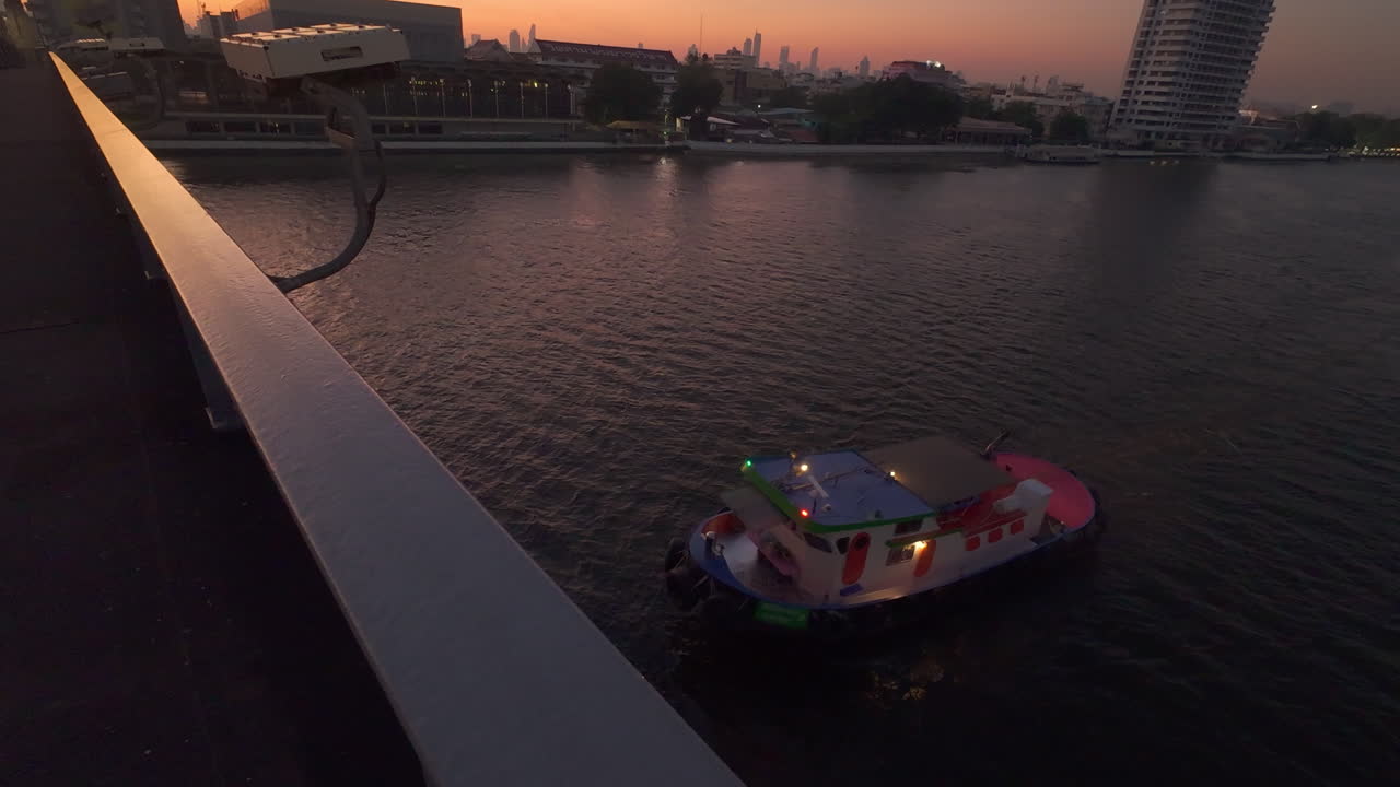 해가 뜨는 동안 도시 풍경을 가로질러 흐르는 조용한 강, 강 위와 다리 아래로 큰 배를 운반하는 견인선을 특징으로합니다.