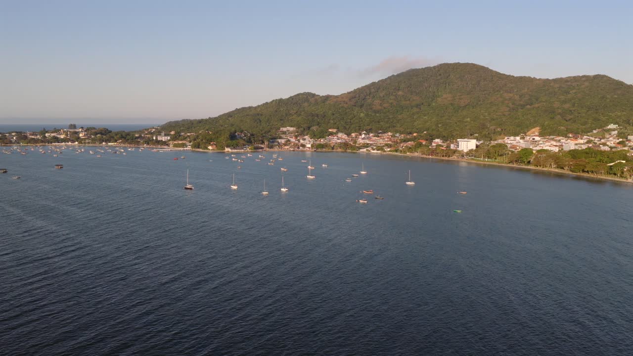 Zoom-in aerial of Penha coastline with anchored fishing boats, hills, and cityscape in Santa Catarina, Brazil