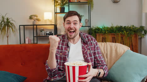 happy man eating popcorn watching home tv shouting celebrating success winning goal game at home