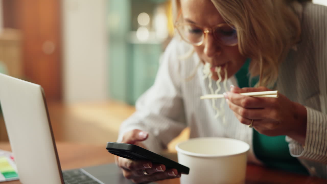 Woman Eating Noodles with Chopsticks While Using Smartphone at Desk