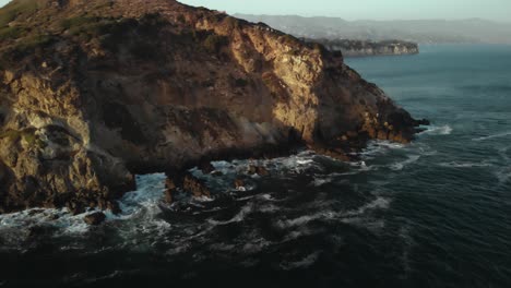 an aerial shot of the point dume cliffs in malibu in california as the waves crash against the rocks in the evening as the vibrant sun sets