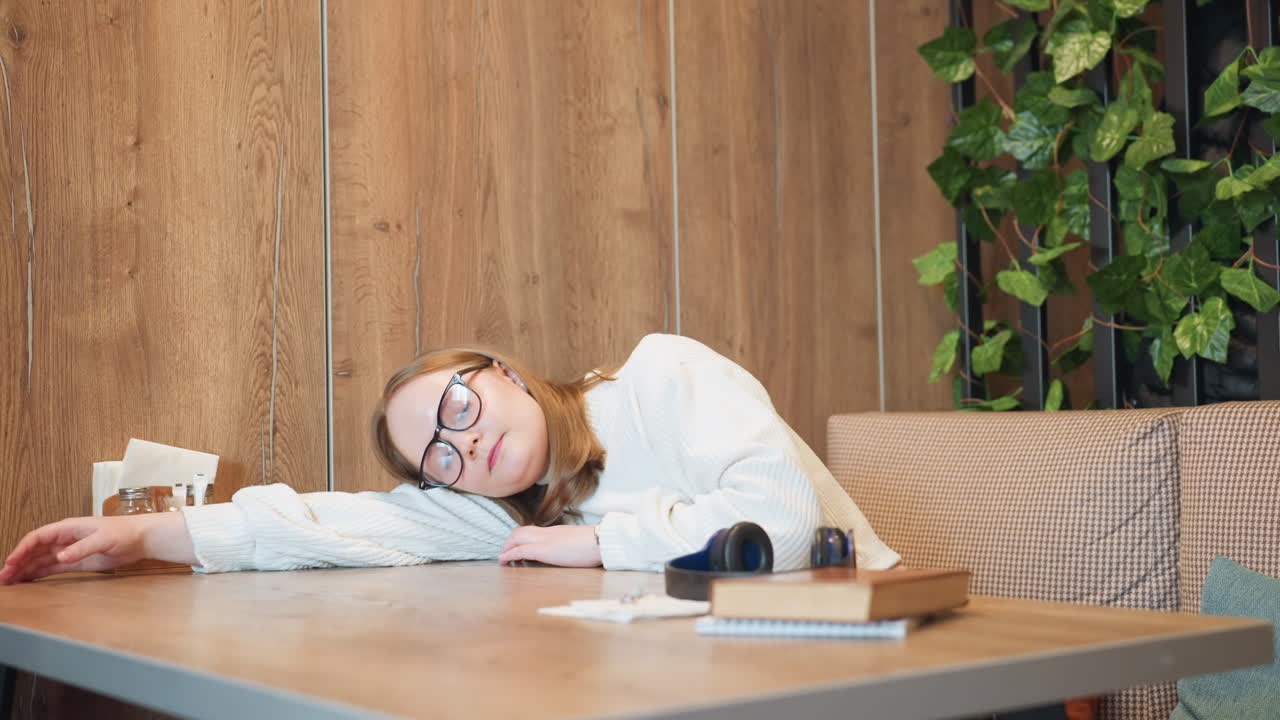 Tired woman in glasses and white sweater removes headset with eyes closed, preparing to rest her head on wooden table with sketches and book in peaceful cafe setting with green decor