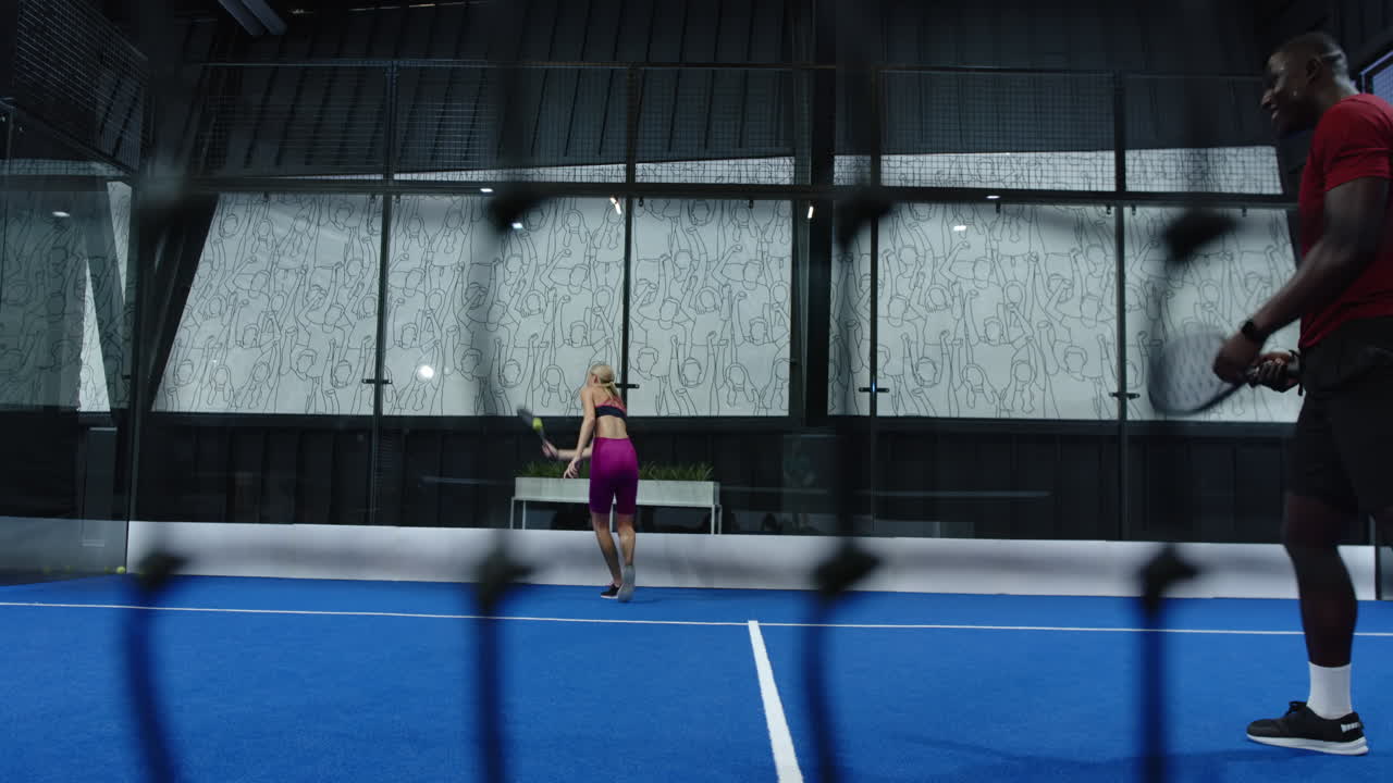 Playing padel tennis, woman in pink outfit hitting ball on blue indoor court