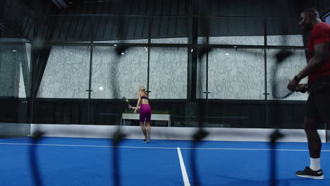 playing padel tennis, woman in pink outfit hitting ball on blue indoor court