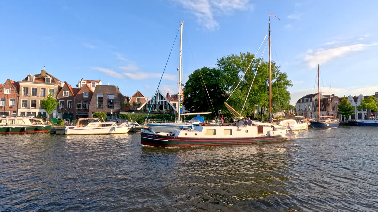 Sailing boat glides along canal, passing waterfront houses under bright summer sunlight, steady camera