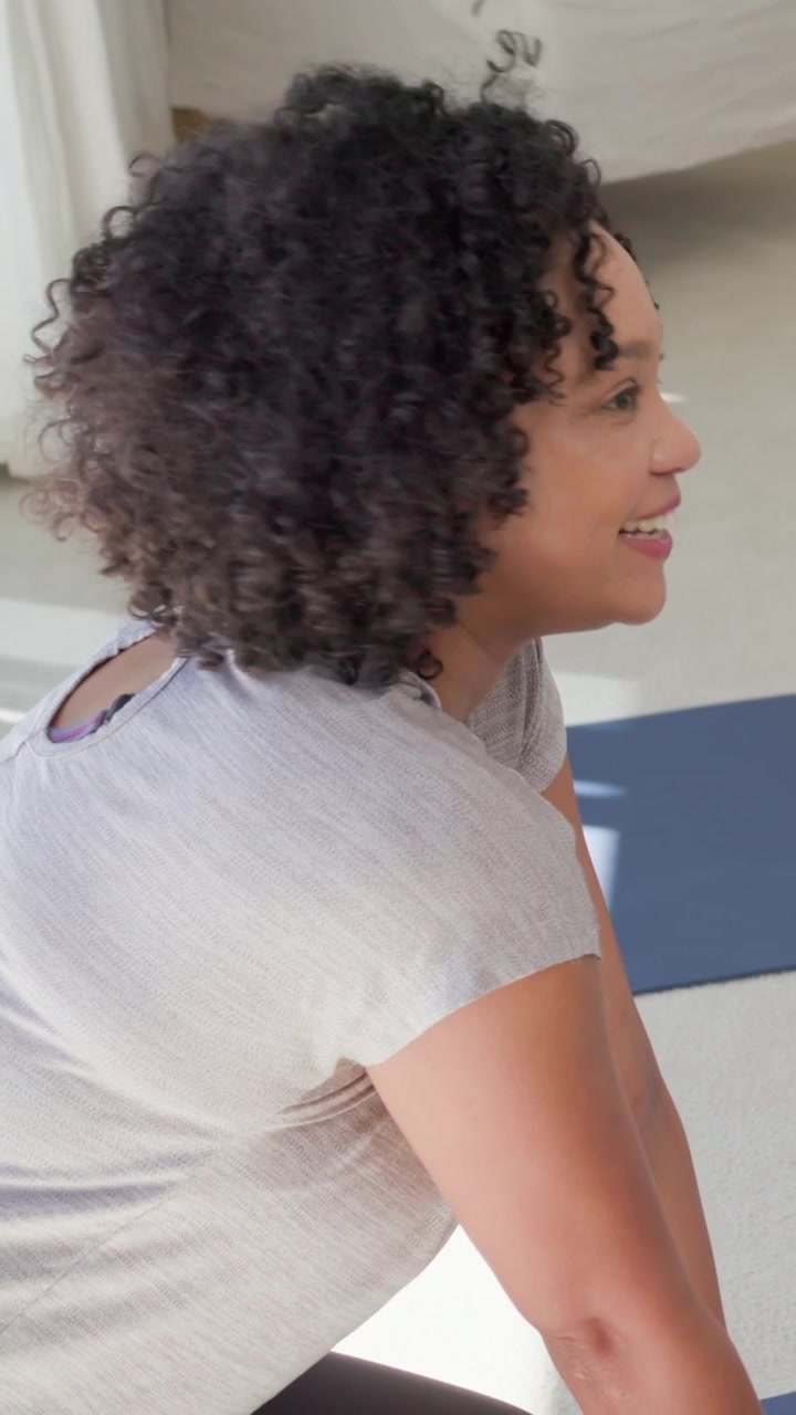 Vertical video: Turning toward light, woman smiling on blue mat in bedroom, gray top strap visible