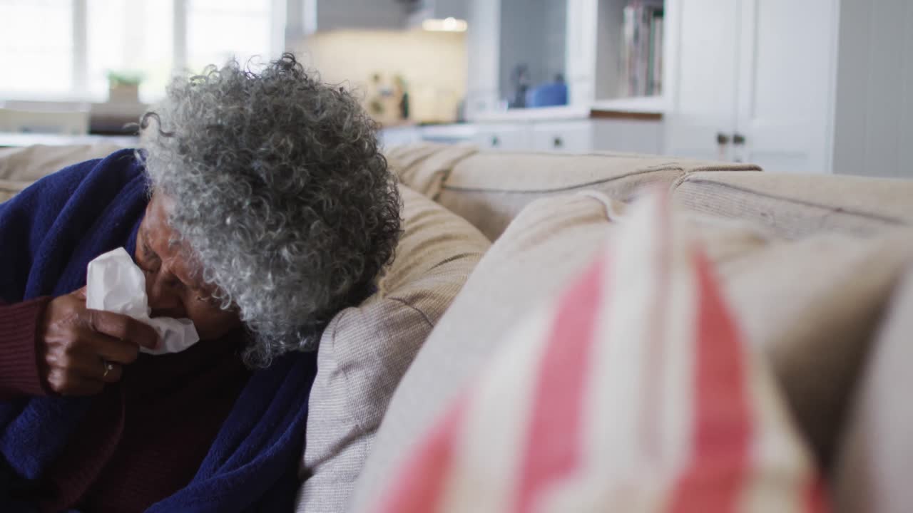 Sick senior african american woman covered with blanket sneezing while lying on the couch at home