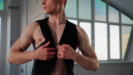 muscular young man before wall size mirror swings black jacket over raised arms, revealing nordic tattooed chest and fit torso while morning city light fills spacious loft