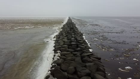 low angle aerial drone view of a long stone mole breaking down a sheet of ice in winter