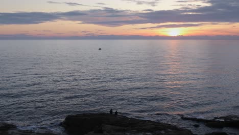 Amantes-Sentados-En-Las-Rocas-Frente-Al-Mar-Durante-La-Puesta-De-Sol-En-Un-Día-Claro-De-Invierno-Y-Un-Pequeño-Barco-De-Pesca-En-El-Mar