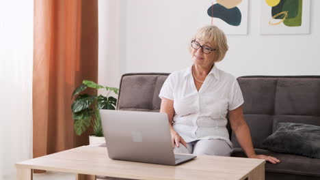 happy senior woman sitting on sofa in living room greeting and talking on video call on modern laptop 2