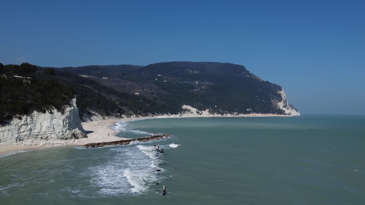 Spiaggia sabbiosa con rocce, scogliere, verdi colline boscose e onde del mare Adriatico, sotto un cielo sereno, all'inizio della primavera, una coppia passeggia sulla spiaggia.