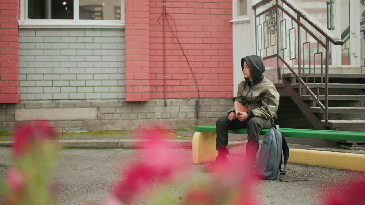 Student in hooded jacket seated on green bench holding and tapping book with right hand, backpack placed nearby, brick wall and staircase behind, blurred flower slightly visible in foreground