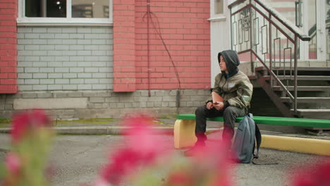 student in hooded jacket seated on green bench holding and tapping book with right hand, backpack placed nearby, brick wall and staircase behind, blurred flower slightly visible in foreground