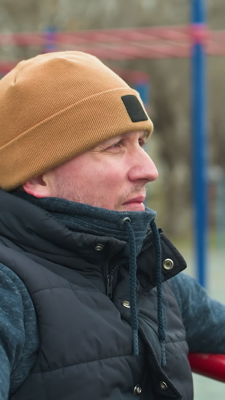 Close-up of a coach sitting with hands resting on workout equipment, looking tired, as he shakes his head, in the blurred background, a boy stands near blue poles