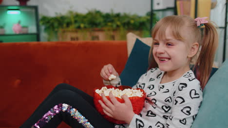 funny children girl watching comedy video film on tv, eating popcorn on comfortable sofa at home