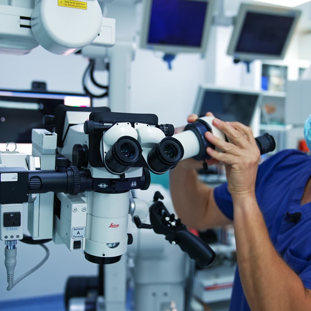 Adult male doctor in blue mask and uniform arranges the equipment. Man adjusting the microscope before the operation. Blurred backdrop