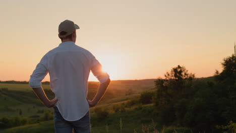 Un-Joven-Agricultor-Confiado-Se-Encuentra-Frente-A-Un-Paisaje-Pintoresco-Mientras-Se-Pone-El-Sol.-Vista-Desde-Atrás