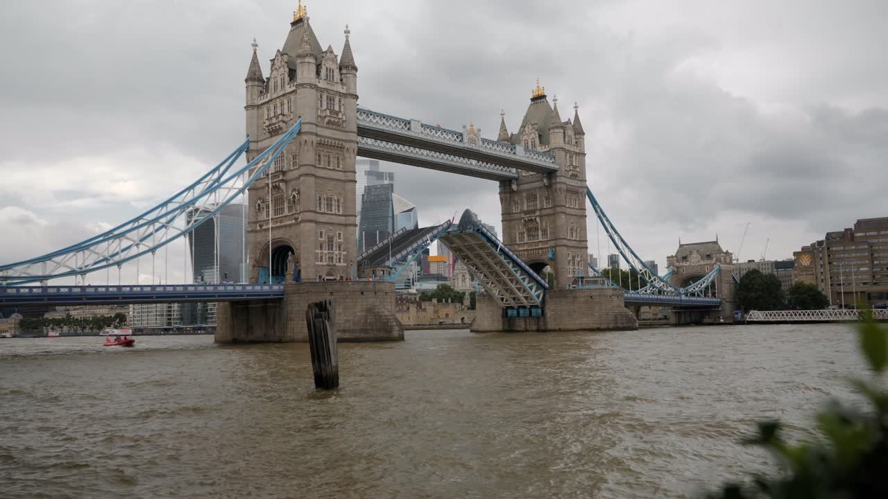 Static shot of London's iconic Tower Bridge with its bascules raised, allowing river traffic passage on the Thames under a cloudy sky.