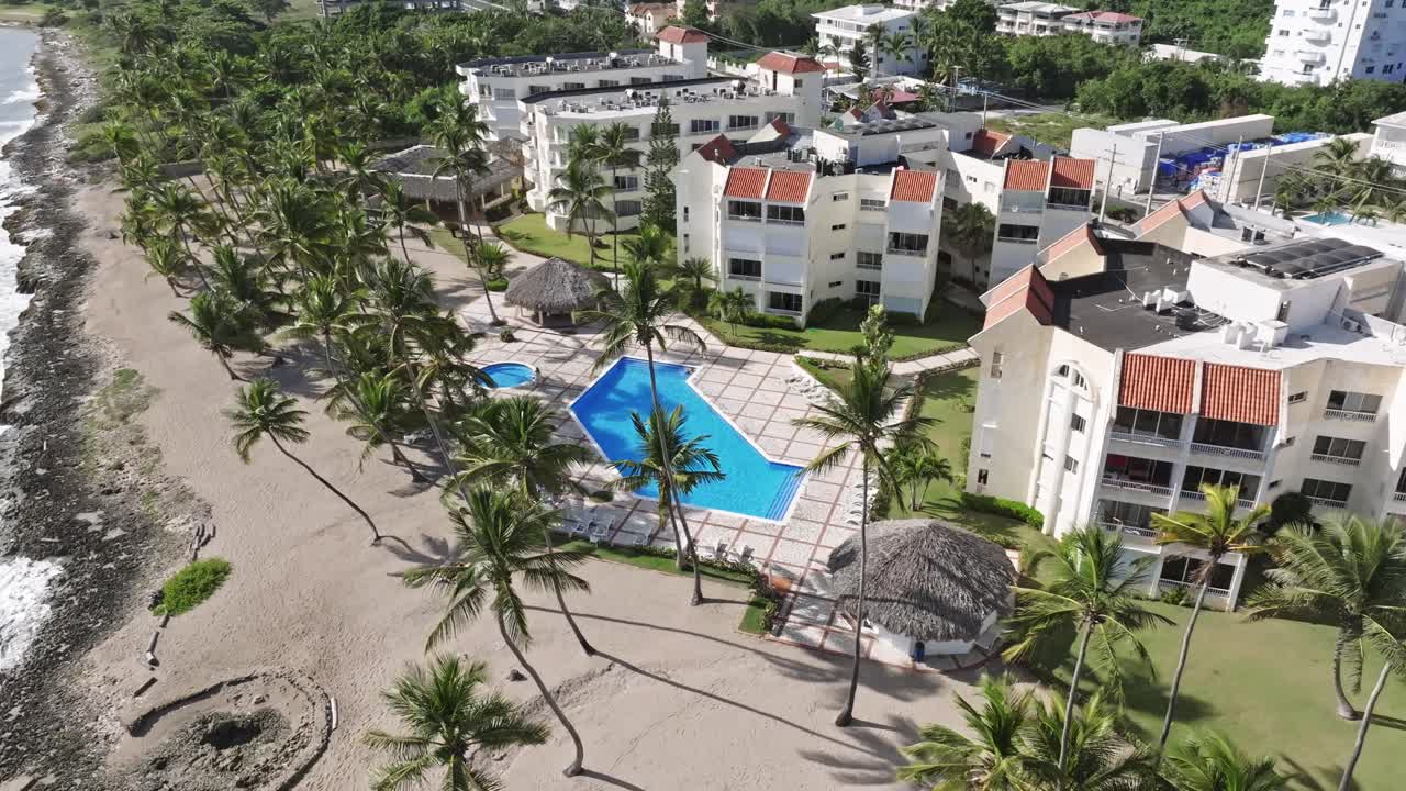 Toma aérea a vista de pájaro de un apartahotel con piscina y palmeras durante un día soleado en Juan Dolio, República Dominicana - Vuelo orbital cenital.