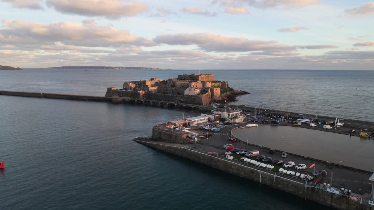 Volo sopra il porto di St Peter Port, Guernsey, sorvolando l'Albert Pier e lo stagno delle barche a vela in miniatura verso il Castello Cornet durante l'ora d'oro, con viste lontane su Sark e il mare blu calmo.