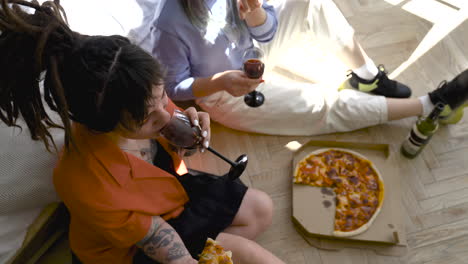 top view of two happy girls drinking wine and sharing pizza while laughing sitting on the floor at home