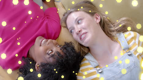 animation of flashing yellow light spots over two happy diverse teenage girls lying on couch talking