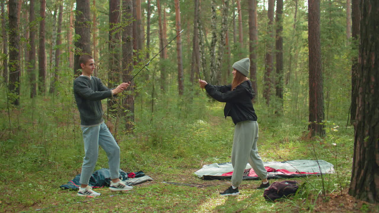 Young couple mimic karate moves with tent poles while camping in forest, surrounded by camping gear scattered on grass, enjoying playful moment together in natural outdoor setting filled with trees
