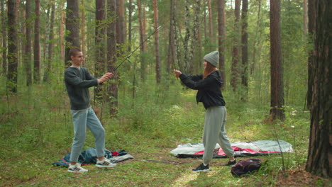 young couple mimic karate moves with tent poles while camping in forest, surrounded by camping gear scattered on grass, enjoying playful moment together in natural outdoor setting filled with trees