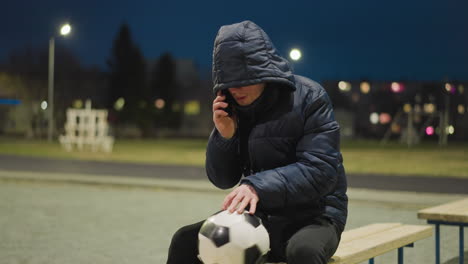 a man sits on a bench in a dimly lit outdoor setting, with a soccer ball between his legs, as he answers a call with his phone with his right hand while gently rotating the ball with his left hand