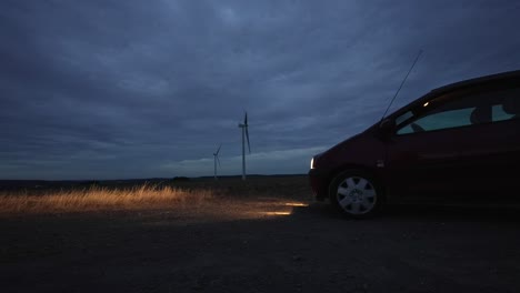 Lapso-De-Tiempo-Del-Día-A-La-Noche-En-Un-Campo-Con-Turbinas-Eólicas,-Faros-De-Automóviles-Iluminan-El-Camino