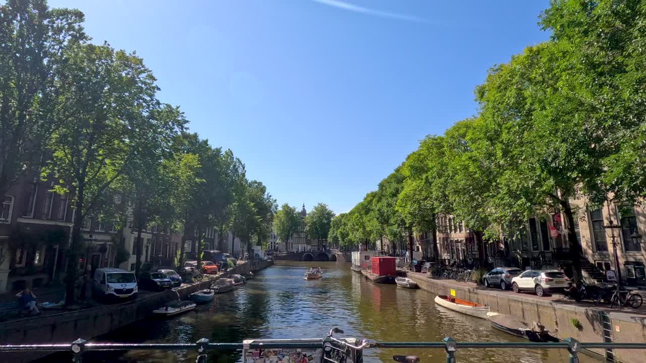 Camera glides over Haarlem canal, capturing boats, tree-lined banks, historic architecture, and bright sunlight