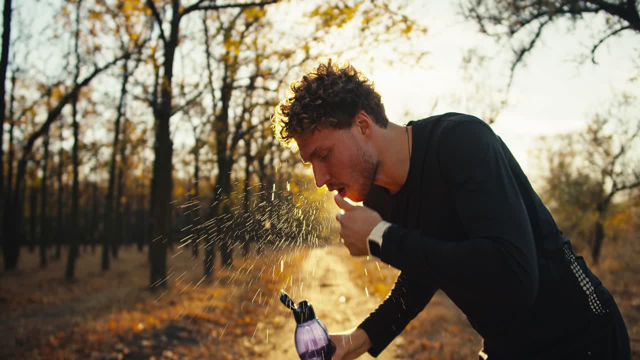 A happy and tired man with curly hair in a black sports uniform pours water from a sports bottle on his face and refreshes himself after a hard jog in an autumn park in the morning