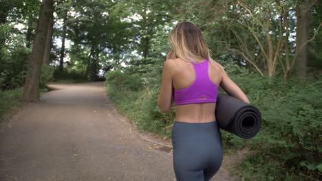 A-tall-blonde-woman-in-exercise-clothing-talks-on-her-phone-as-she-walks-down-a-wooded-park-path-carrying-a-yoga-mat