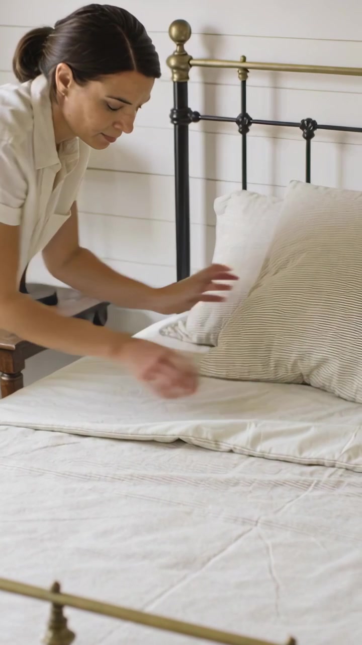 Video vertical: Mujer en blusa inclinándose, arreglando la cama ahuecando una almohada en el dormitorio, con espacio para texto