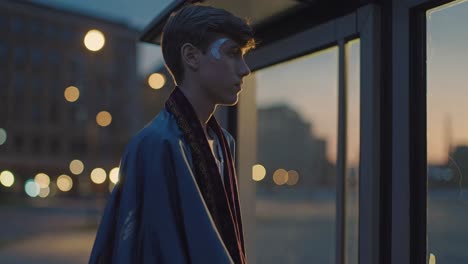 young man with a cape and a crescent moon painted on his face is waiting at a bus stop at dusk, looking outside, with city lights blurred in the background