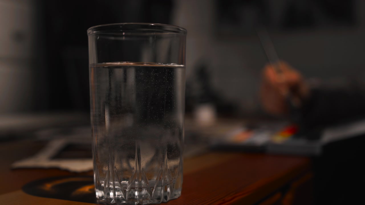 Glas vand på træbord med sløret hånd i baggrunden, bløde refleksioner og varm stemning