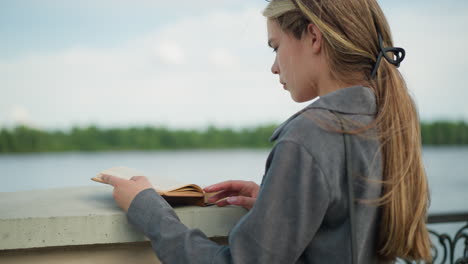 lady in grey shirt reading a book while resting on a fence, the wind gently blows the pages as she remains focused, with a tranquil water and greenery background