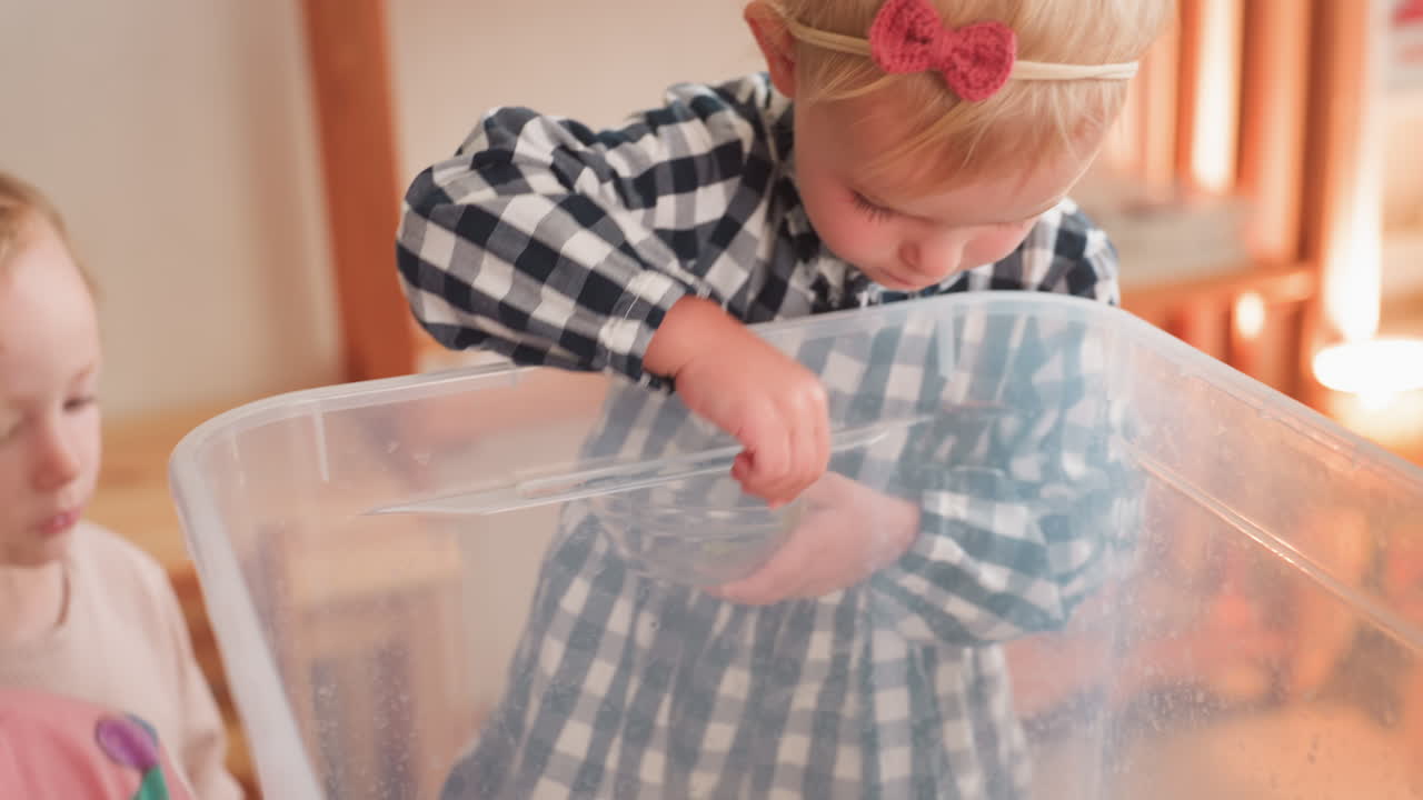 Children leaning over transparent rubber container, observing contents inside with curiosity while one child holds small bowl, creating tender educational moment of focus and attention