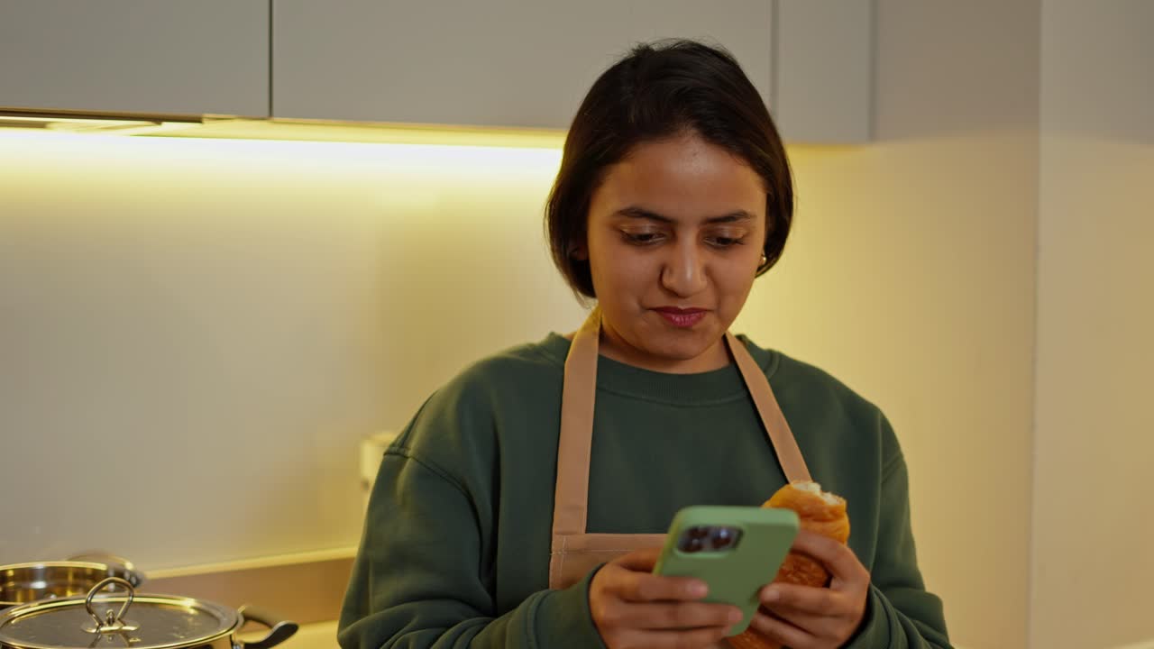 feliz chica morena en un suéter verde oscuro y delantal beige está escribiendo a sus amigos en el teléfono durante su descanso y almuerzo con un croissant en la cocina en un apartamento moderno
