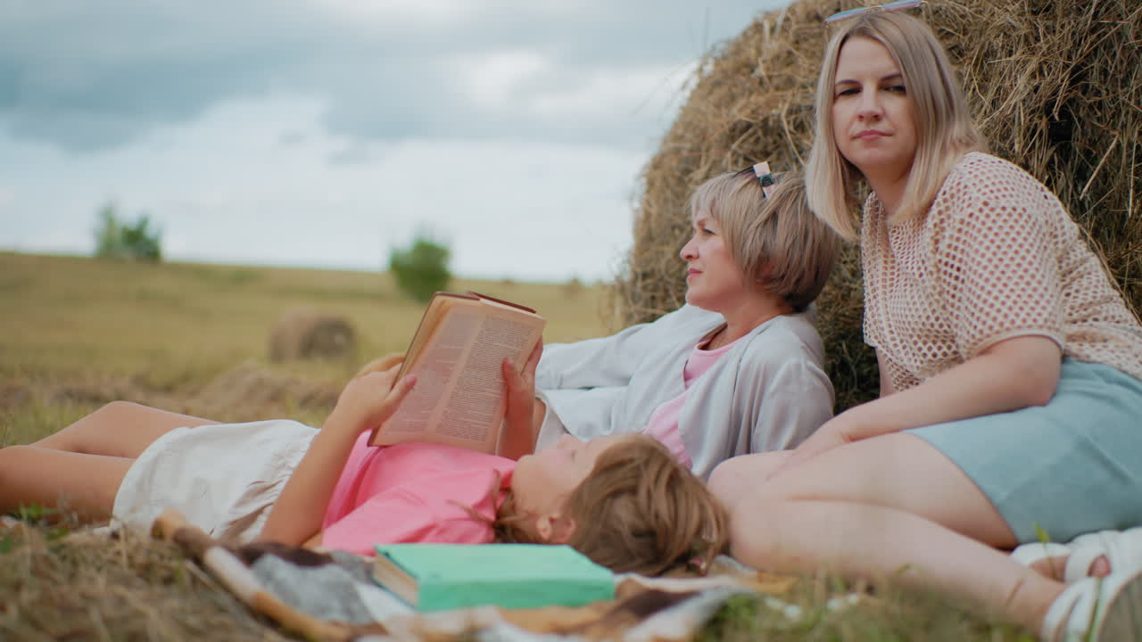 семья наслаждается спокойным пикником на одеяле на обширной сельскохозяйственной земле, мать и дочь расслабляются с книгами, собака отдыхает рядом с ними, прекрасный момент семейной связи на открытом воздухе в природе